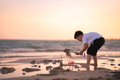 一个小男孩在夕阳下的海边的沙滩上挖沙子