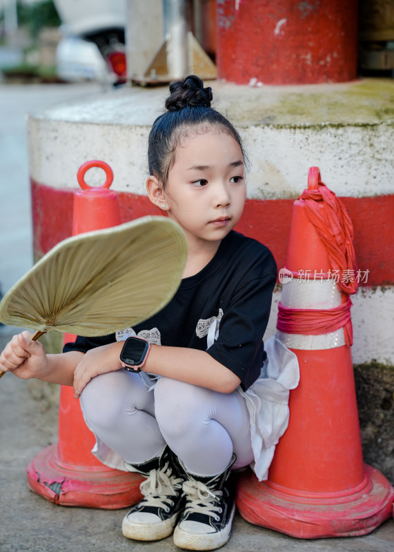 小女孩蹲在交通锥旁手持扇子