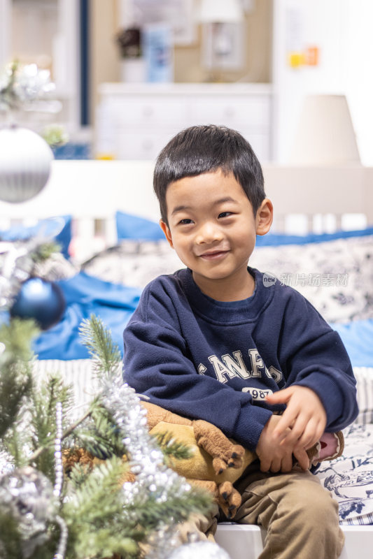 一个小男孩抱着毛绒玩具坐在圣诞树旁