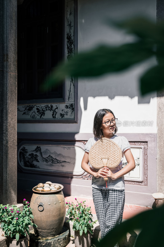 手持草编扇的女孩站在中式古建庭院内