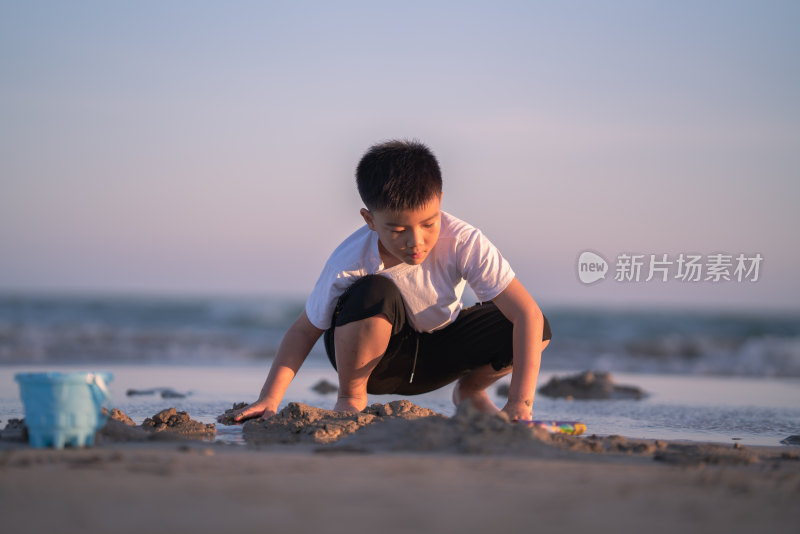 一个小男孩在夕阳下的海边的沙滩上挖沙子